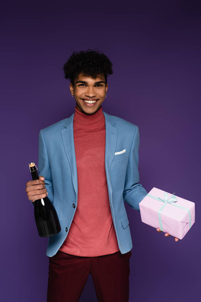 szczęśliwy Afrykański Amerykanin w niebieskiej marynarce trzyma zapakowany prezent i butelkę szampana na fioletowym - Zdjęcie, obraz