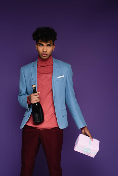 fiatal afro-amerikai férfi kék blézerben, kezében csomagolva ajándék és üveg pezsgő lila - Fotó, kép