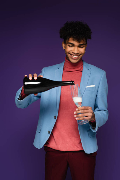 heureux homme afro-américain en blazer bleu verser du champagne dans un verre sur violet - Photo, image