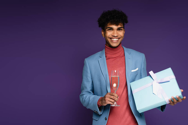 cheerful african american man in blue blazer holding glass of champagne and present on purple - Photo, Image