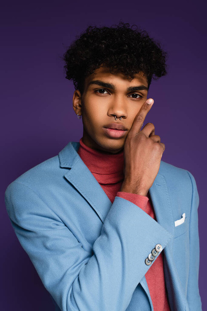 pierced african american man in blue blazer touching cheek on purple - 写真・画像