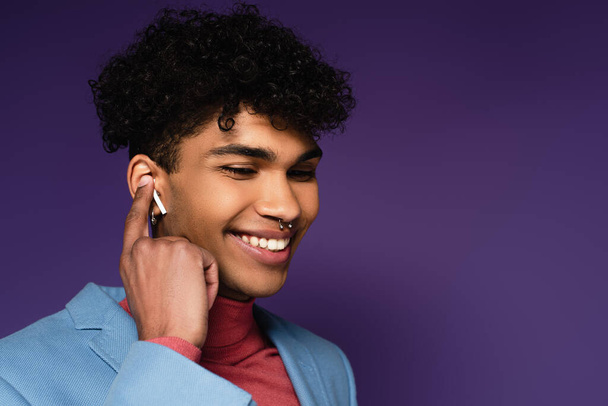 hombre afroamericano feliz ajustando auriculares inalámbricos en púrpura - Foto, imagen