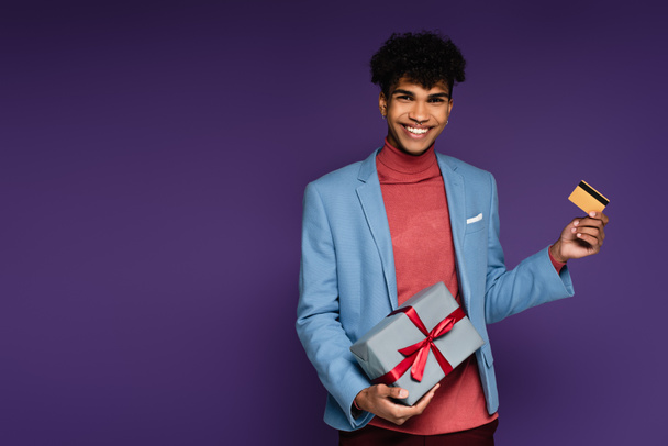 cheerful african american man in blue blazer holding credit card and present on purple - Photo, Image