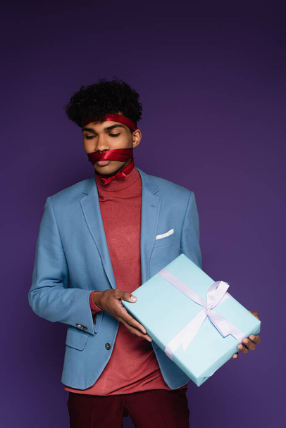 african american man tied with red ribbon holding present on purple  - Photo, Image