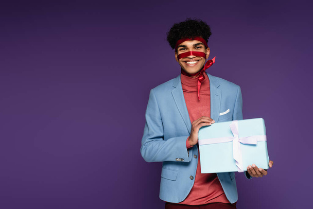 радісний афроамериканський чоловік пов'язаний з червоною стрічкою тримає подарунок на фіолетовому
  - Фото, зображення