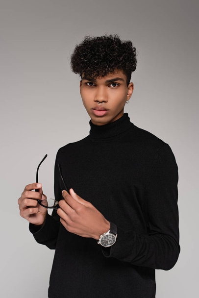 young african american man in black turtleneck sweater holding glasses and posing isolated on grey - Фото, изображение