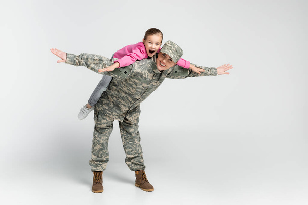 Erstaunte Mädchen huckepack auf Mutter in Camouflage auf grauem Hintergrund  - Foto, Bild