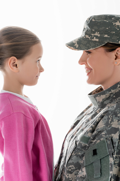Vista lateral del niño sonriente mirando a la madre en uniforme militar aislado en blanco  - Foto, Imagen