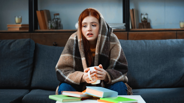 Red haired girl wrapped in blanket holding cup near books on blurred foreground  - 写真・画像