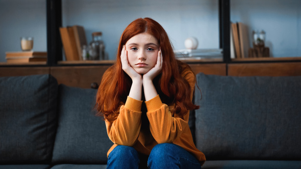 Red haired teenager looking at camera while sitting on couch at home  - Photo, Image