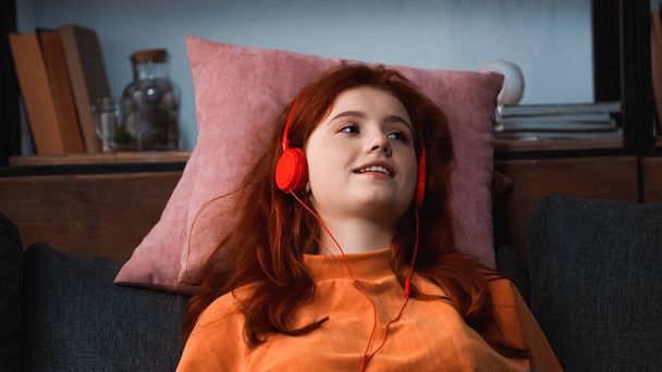 Chica sonriente escuchando música en auriculares en almohadas  - Foto, Imagen