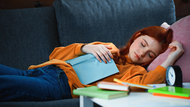 Κοκκινομάλλα κορίτσι κοιμάται κοντά στο βιβλίο και ξυπνητήρι σε θολή πρώτο πλάνο  - Φωτογραφία, εικόνα