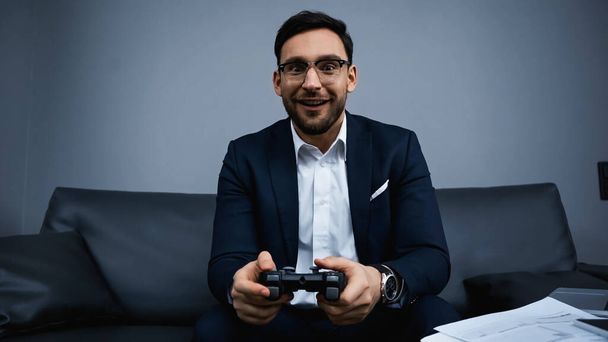 KYIV, UKRAINE - FEBRUARY 13, 2020: Smiling businessman playing video game near papers in hotel  - Fotoğraf, Görsel