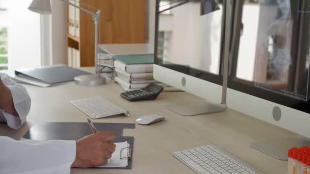 Links van middelbare leeftijd mannelijke Aziatische arts zitten aan het bureau in zijn kantoor, kijken naar computerschermen, praten over de telefoon, terwijl het maken van aantekeningen in patiëntenkaart - Video