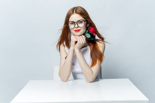 jolie femme rousse assise à la table avec fleur de rose dans les mains glamour rouge lèvres modèle - Photo, image