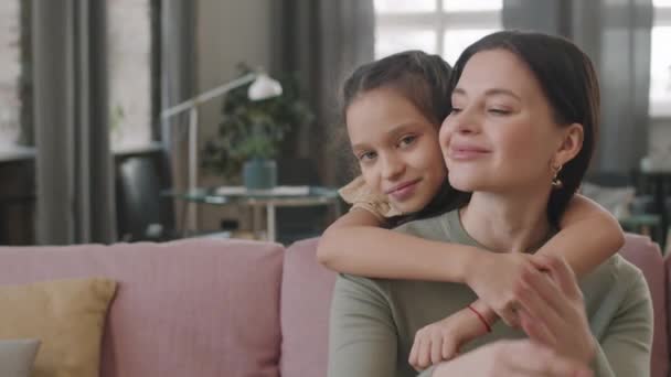 Slow-motion panning medium close-up portret van mooi klein meisje knuffelen haar moeder, beide kijken naar camera glimlachen, thuis blijven in lichte gezellige woonkamer - Video