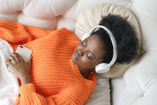 African millennial woman in orange sweater wearing wireless headphones, relaxes enjoying her favorite music, resting with closed eyes, lying on couch in living room. Girl spend lazy weekend at home. - Photo, Image