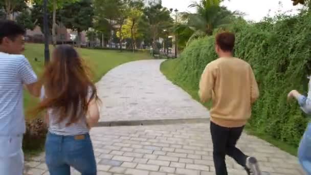 Brede opname van twee jonge Aziatische mannen en hun twee vriendinnen die wegrennen in het park, dan stoppen, omdraaien, naar de camera kijken en lachen - Video