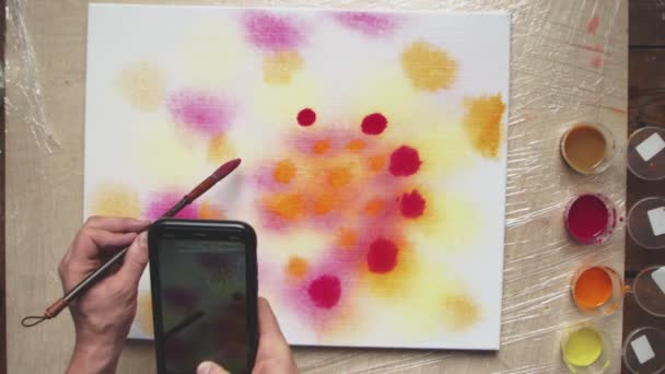 Κάτοψη του αρσενικού καλλιτέχνη ζωγραφίζει Pastel tie dye ή Pastel εικόνα στροβιλισμού με κόκκινους κύκλους σε βρεγμένο καμβά και καταγράφει βίντεο από αυτό στο τηλέφωνό του. Ρευστή τέχνη. - Πλάνα, βίντεο