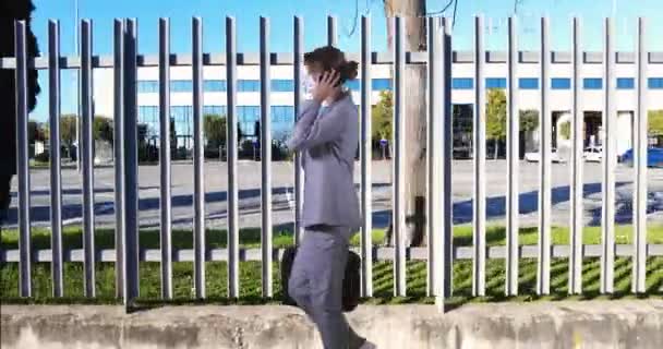 Nuori liikenainen, jolla on IIR-naamio, kävelee toimistorakennuksen metallisen aidan taustalla vastaamassa puhelimeen - Materiaali, video