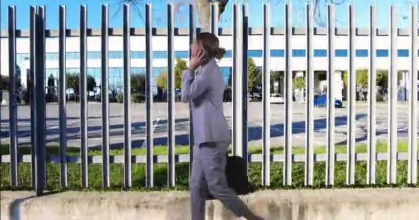 Νεαρή επιχειρηματίας περπατά στο βάθος ενός μεταλλικού φράχτη ενός κτιρίου γραφείων απαντώντας στο τηλέφωνο - Πλάνα, βίντεο