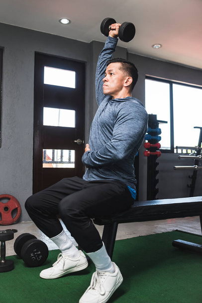 Egy sportos ázsiai fickó pulóverben ül a feje fölött, fél kar tricepsz hosszabbítóval. Edzés és tréning tricepsz. Szabadtéri edzőterem beállítása. - Fotó, kép