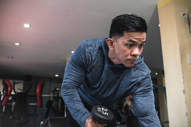 Un hombre asiático apático y aparentemente desmotivado hace sobornos de tríceps de pesa en un banco en un gimnasio. Ejercicio y entrenamiento de tríceps y brazos. - Foto, imagen