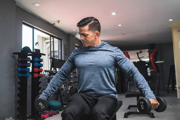 A fit asian man in a sweatshirt does seated side lateral raises. Shoulder workout and training. Open air gym setting. - Foto, Imagem