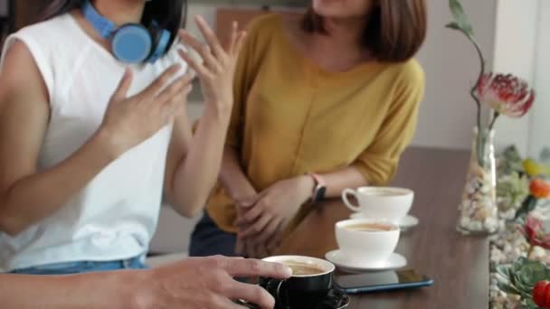Két ázsiai nő és kevert bőrű férfi ül együtt a kávézóban, beszélgetnek és cappuccinót isznak. - Felvétel, videó