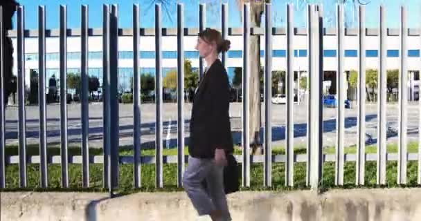 Jonge zakenvrouw wandelen en denken op de achtergrond van een metalen hek van een kantoorgebouw - Video