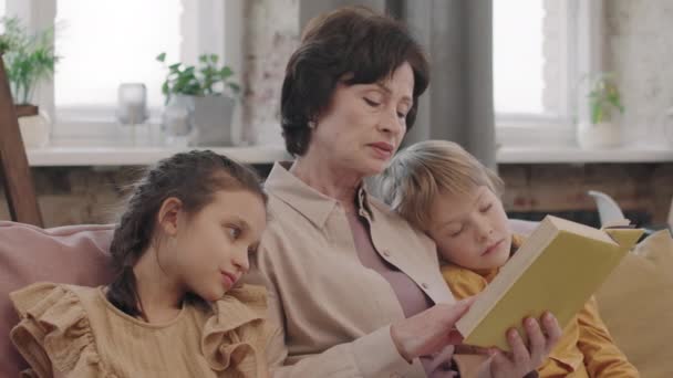 Panning medium close up with slowmo of loving γιαγιά ανάγνωση χάρτινο βιβλίο δυνατά στα μικρά εγγόνια της που βρίσκονται στους ώμους της στον καναπέ στο σαλόνι - Πλάνα, βίντεο