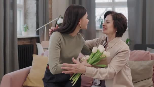 Mittlere Aufnahme einer attraktiven erwachsenen Frau, die ihrer Mutter eine Geschenkbox und einen Strauß weißer Tulpen schenkt, zu Hause gemeinsam Geburtstag feiert, lächelt und umarmt - Filmmaterial, Video