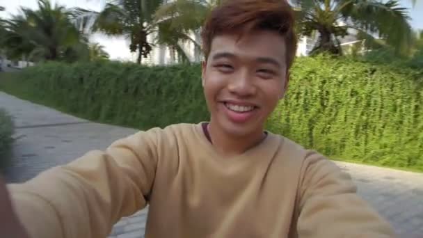 Asian teenager holding camera, making selfie and smiling while his girlfriend standing and watching him - Footage, Video