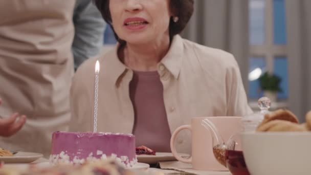 Zwolnione tempo zbliżenie przystojnej dojrzałej kobiety uśmiechającej się zdmuchując świeczkę na torcie urodzinowym przed rodziną siedzącą przy stole obiadowym - Materiał filmowy, wideo