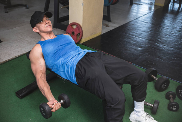 A fit asian man in a blue tank top does lying dumbbell curl on a bench. Challenging bicep isolation exercise and workout at the gym. - Photo, Image
