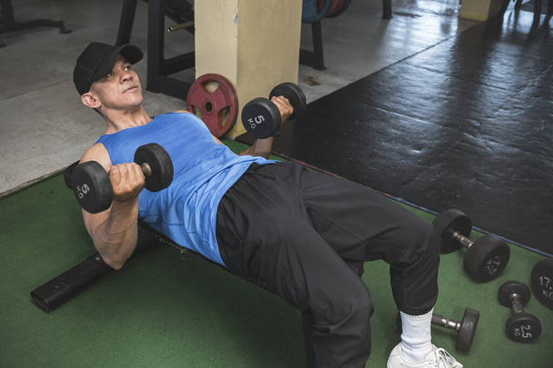A fit asian man in a blue tank top does lying dumbbell curl on a bench. Challenging bicep isolation exercise and workout at the gym. - Photo, Image