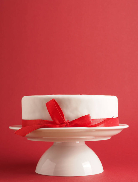 Biały tort weselny lub urodzinowy z czerwoną kokardą na białym stoisku z ciastami. Czerwone tło z miejscem na tekst. - Zdjęcie, obraz