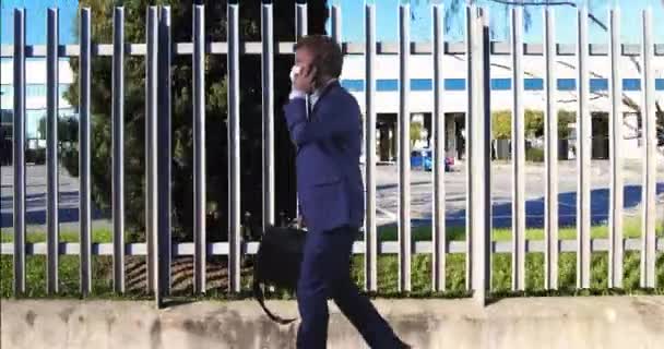ffp2マスクを持つ若いビジネスマンが電話に応答するオフィスビルの金属フェンスの背景を歩く - 映像、動画