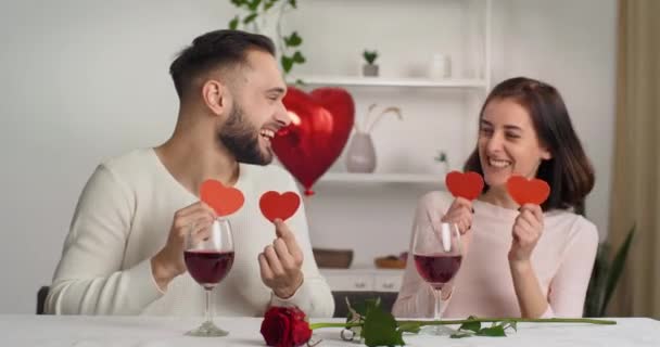 Coppia barbuta giovane uomo caucasico e bruna ragazza sinceramente ridendo divertirsi insieme a casa sulla celebrazione dell'anniversario di San Valentino applicando ai loro occhi cuori rossi in posa per la fotocamera - Filmati, video