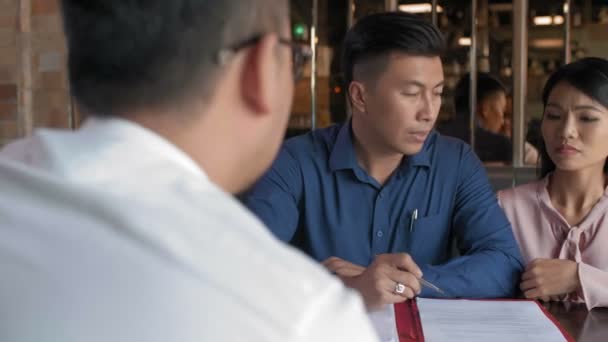 Indietro vista di coppia asiatica seduta a tavola e discutere documento con il loro avvocato seduto di fronte a loro - Filmati, video