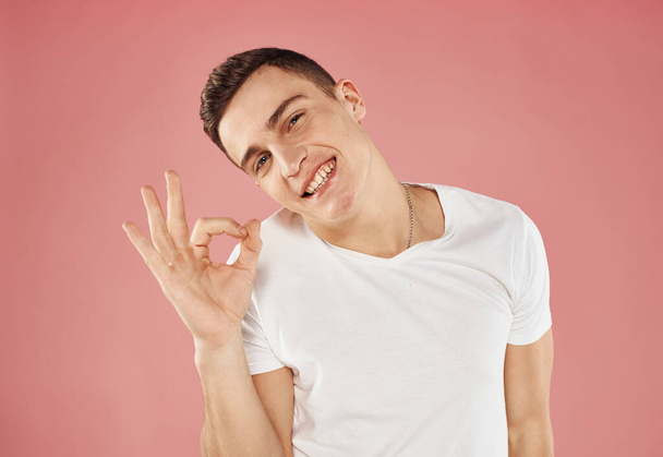 Leuke man en wit t-shirt hand gebaar emoties roze achtergrond - Foto, afbeelding
