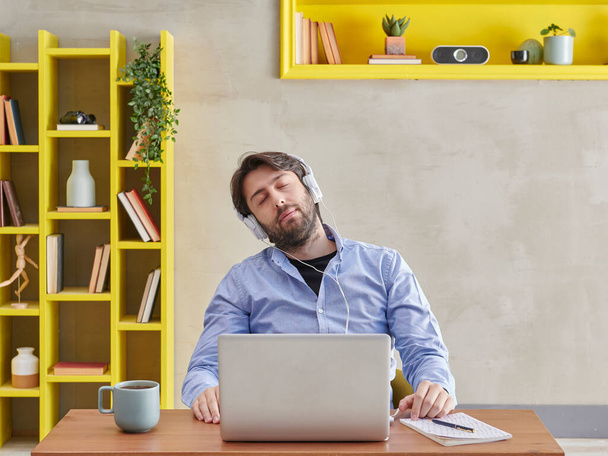 Άντρας σε μπλε πουκάμισο αποκοιμιέται σε βίντεο μιλώντας, γκρι φόντο στον τοίχο και κίτρινο ράφι, καφέ laptop στυλ. - Φωτογραφία, εικόνα