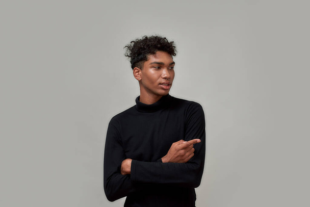 Cool young guy with piercing dressed in black looking and pointing at right side, presenting something, standing isolated over gray background - 写真・画像