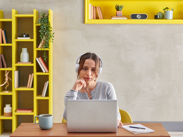 Vrouw luistert muziek in video praten, op zoek naar het scherm, grijze muur achtergrond en gele boekenplank, koffie laptop stijl. - Foto, afbeelding