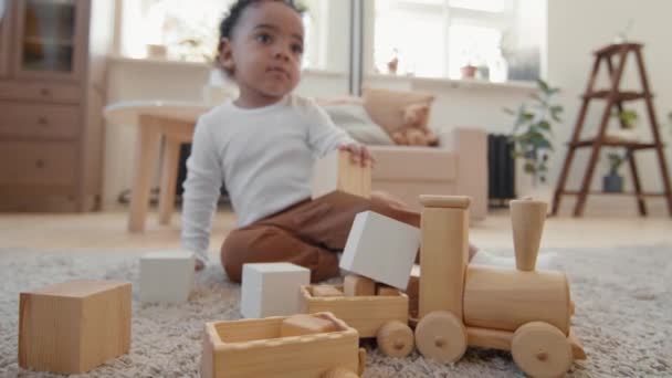 Handheld tracking met slowmo van schattige peuter jongen zittend op tapijt in gezellige woonkamer en spelen met houten trein speelgoed en bouwstenen - Video