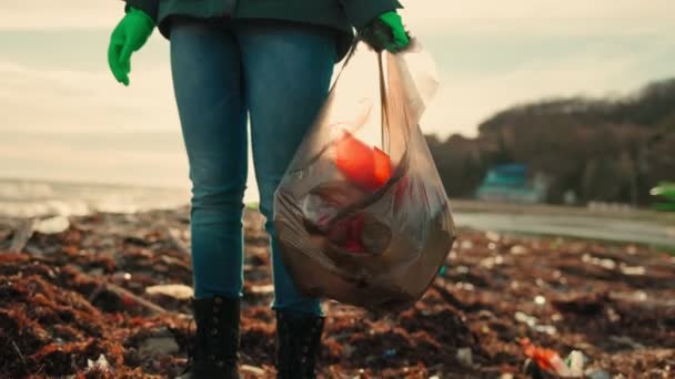 A young female volunteer wearing rubber gloves, holding a garbage bag. Camera movement from bottom to top. Sunset. Slow motion. Concept of Earth Day. - Footage, Video