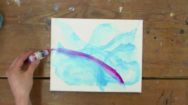 Vista superior de la artista pinta un cuadro abstracto, ella pinta formas abstractas con pintura púrpura sobre lienzo húmedo con cuadro azul y utiliza pincel para distribuirlo - Metraje, vídeo
