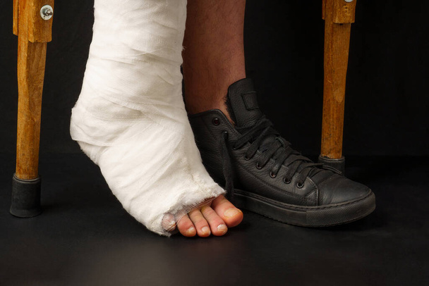 un hombre con un tobillo roto en un yeso en su pierna, camina sobre muletas, sobre un fondo negro - Foto, Imagen