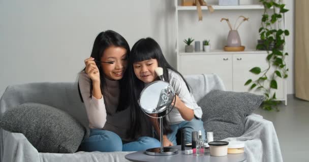Little daughter and loving mother asian family two girls doing makeup beauty procedures at home sitting on couch looking in mirror use cosmetics mascara and blush take care of skin spend time together - Footage, Video