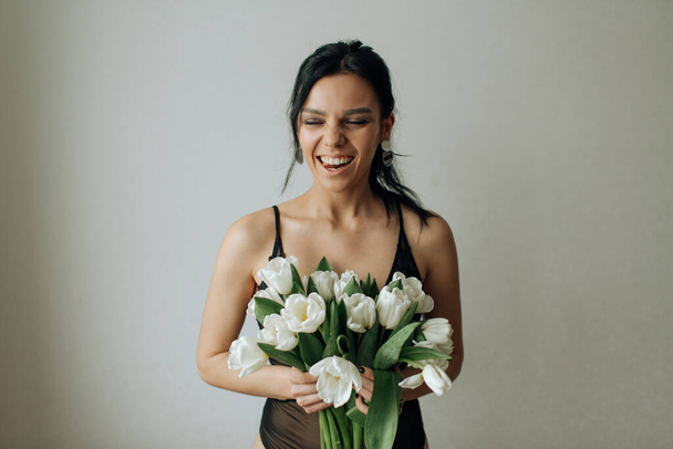 Beautiful brunette girl in a black bodysuit laughing holding a bouquet of white tulips in her hands - Foto, immagini